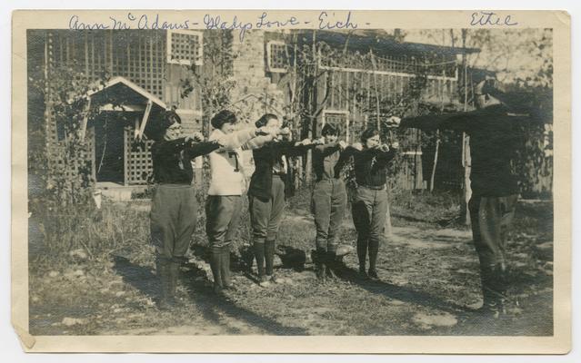 Ann McAdams, Gladys Lowe, Eich, Ethel, setting up exercises [standing in a row with arms stretched out while a man stands facing them with his arms stretched out], camping on Kentucky River, 1919-1923.  A cabin is in the background