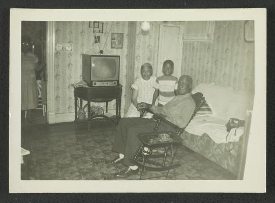 Unidentified elderly black man and woman, seated, with child standing between them; unidentified woman standing in doorway on left side