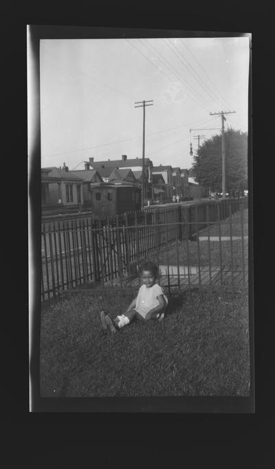 Negative of an unidentified child sitting in a fenced-in yard