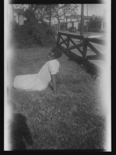 Negative of unidentified woman sitting in a fenced-in yard