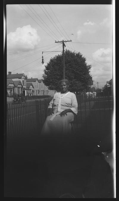 Negative of an unidentified elderly woman sitting on a hair in a fenced-in yard