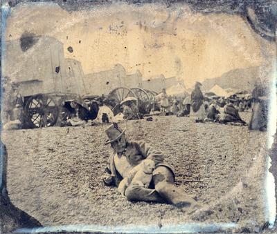 Unidentified man lounging in field, wagons and tents in background