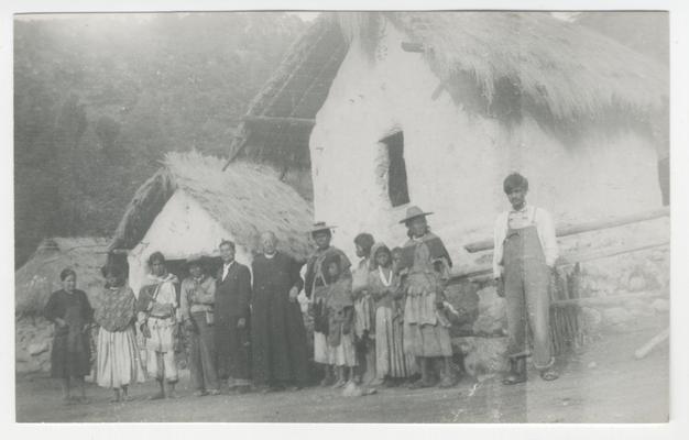 A group of thirteen adults and children (one a child in the arms of another person) stand in front of three whitewashed, stone buildings, each with a thatched roof in Los Gatos, Jalisco, Mexico.  At least ten of the people are probably Wixárika.  At least two of the people are Catholic priests