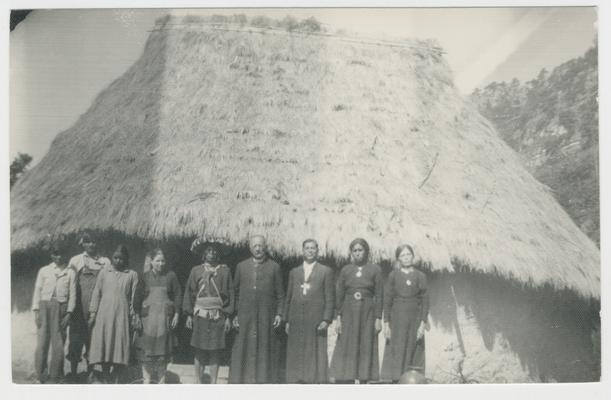 A group of nine adults and young adults stand in front of a structure with whitewashed walls and a thatched roof
The majority of the people are probably Wixárika. Two of the people are Catholic priests