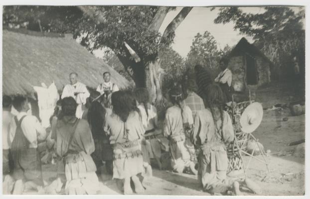 A large group of people kneel on the ground with their backs to the photographer, probably in Zalatita, Jalisco, Mexico. Facing them and the photographer are two standing adults. The entire group is kneeling and standing in front of a structure with a thatched roof.  The majority of the kneeling people are probably Wixárika.  The two standing people are Catholic priests. In the background on the right is a tree, a standing person with arms folded, and another structure.  A woven wicker structure with a hat on it is also in the foreground