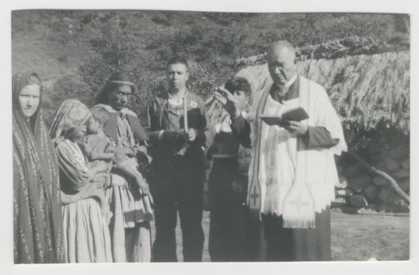Seven adults and children (one child being held by another person) stand in front of a a hill and a stone structure with a thatched roof in Los Pocitos, Jalisco, Mexico.  Most of the people are probably Wixárika. At least one of the adults is a Catholic priest.  The group is probably participating in a Catholic baptismal ceremony