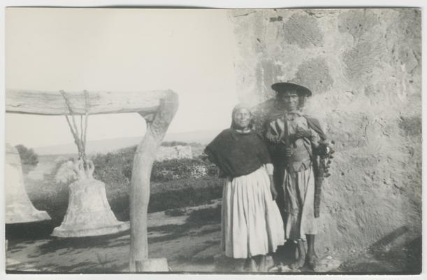 Two people, probably a man and a woman, lean against a stone wall in Zenzompa, Jalisco, Mexico. They have probably just been married. They are probably Wixárika. To the left is a large bell suspended in a stand. In the background is a vista of vegetation and stone structures sloping away from the foreground, with what is probably a large body of water in the distance, probably either the Gulf of California or the Pacific Ocean
