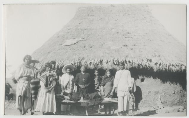 At least nine adults and children (one a child carried by another person) stand in front of a structure with a thatched roof in Los Pocitos, Jalisco, Mexico. A person lies on a cot in the middle of the group. The majority of the people are probably Wixárika. One of them is a Catholic priest. There is probably an animal skin stretched on the thatched roof