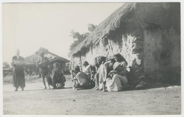 At least ten adults and children stand, sit, or squat in a clearing next to two structures with thatched roofs in Los Gatos, Jalisco, Mexico. The majority of the people are probably Wixárika.  One of the people is probably a Catholic priest. The group is probably learning Catholic catechism being taught by a Wixárika