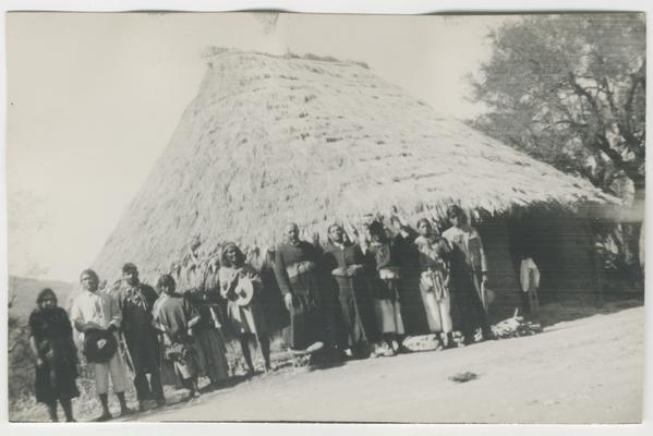 A group of ten adults stand in front of a structure with a thatched roof, possibly a Wixárika temple in Zaymasita, Jalisco, Mexico. The majority of the people are probably Wixárika. Two of the people are Catholic priests