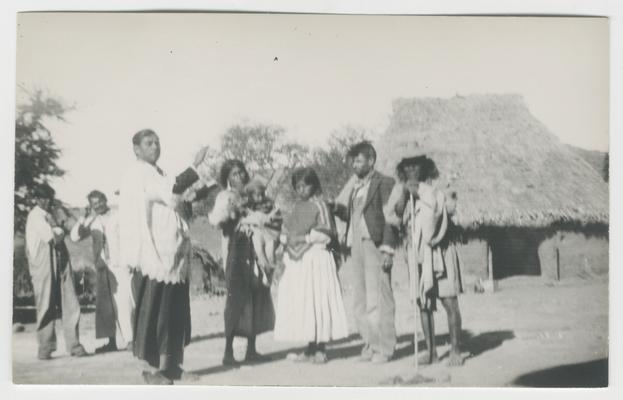 Five adults and children (one a child carried by another person) stand in front of a structure with a thatched roof, probably in Zaymasita, Jalisco, Mexico.  The majority of the people are probably Wixárika. One of the people is a Catholic priest, who stands on the left, looking at the photographer but facing toward the group of four people to the right.  The priest's right hand is outstretched, and he holds a book in his left hand.  Two adults stand in the background.  The people are probably participating in a Catholic ceremony