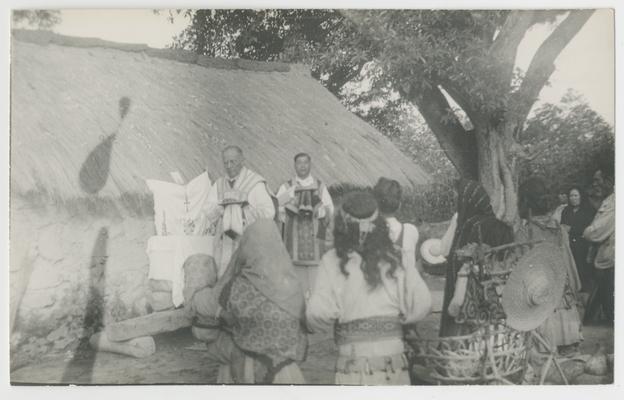 A large group of people kneel on the ground with their backs to the photographer, probably in Zalatita, Jalisco, Mexico. Facing them and the photographer are two standing adults. The entire group is kneeling and standing in front of a structure with a thatched roof. The majority of the kneeling people are probably Wixárika. The two standing people are Catholic priests. In the background on the right is a tree and at least one standing person. A woven wicker structure with a hat on it is also in the foreground