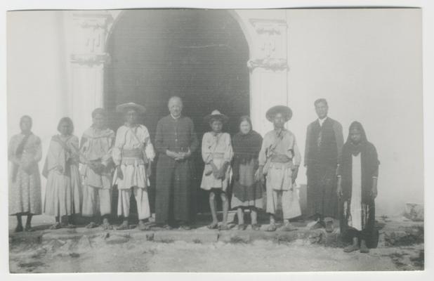 A group of ten adults and children stand in front of an arched doorway in a white wall with plaster decorations, probably a Catholic church building at Zenzompa, Jalisco, Mexico.  At least eight of the people are probably Wixárika.  At least two of the people are Catholic priests