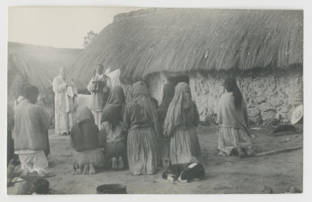 Nine people kneel on the ground with their backs to the photographer. Facing them and the photographer are two standing adults. The entire group is kneeling and standing in front of a whitewashed stone structure with a thatched roof. The majority of the kneeling people are probably Wixárika. The two standing people are Catholic priests. A dog is curled up on the ground behind one of the kneeling people