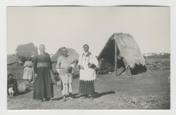 Three adults stand in the foreground with at least two people sitting or standing in the background in front of or next to plaster or wooden structures covered with thatched roofs, probably at San Andrés Cohamiata, Jalisco, Mexico.  One of the adults in the foreground is the Governor and probably Wixárika.  Two of the adults in the foreground are Catholic priests