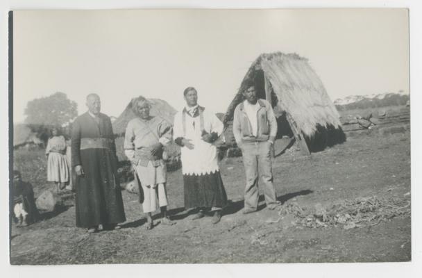 Four adults stand in the foreground with at least two people sitting or standing in the background in front of or next to plaster or wooden structures with thatched roofs, probably in San Andrés Cohamiata, Jalisco, Mexico. One of the people standing is the Governor and the other is a neighbor. Both are probably Wixárika. Two are Catholic priests