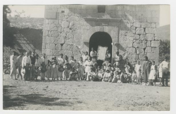 A large group of adults and children stand or sit in front of an arched doorway in a stone structure at Guadalupe Ocotán, Jalisco, Mexico. Two people stand in the doorway, each holding one side of a rectangular framed object. The people are most likely Wixárika. The scene probably documents a celebration of the Catholic feast of Our Holy of Guadalupe on December 12