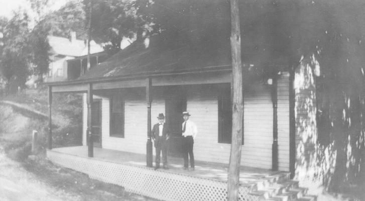 Two men standing on porch of house on dirt road