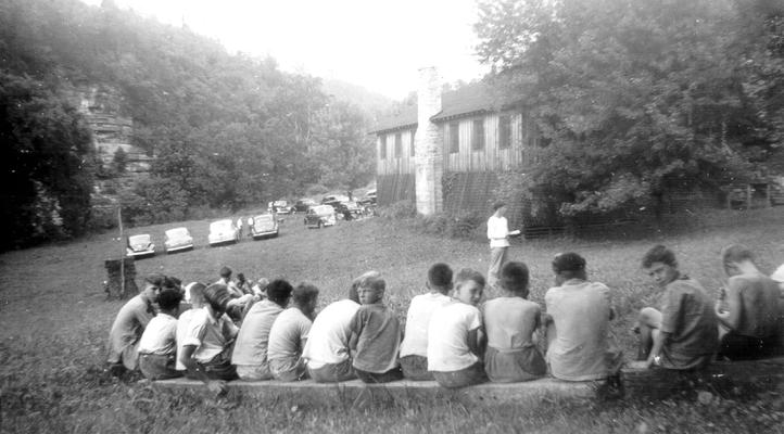 About twenty boys sitting on logs with a teacher up front