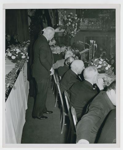 From front row of dais, Secretary Vinson turns to address man standing behind row at New York County War Bond Committee Dinner