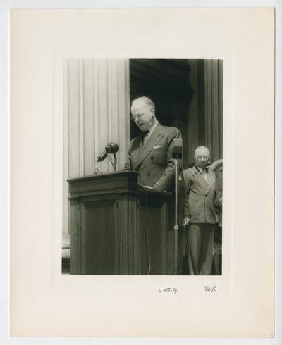 Treasury Secretary John W. Snyder speaks from podium with President Truman standing behind