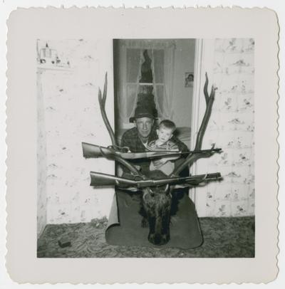 Hunter and child posed behind trophy head with two rifles hanging in antlers