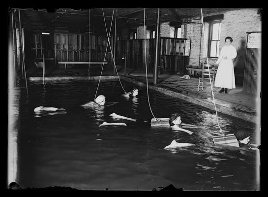 Four women swimming, pool at Lincoln School, Mrs. Stout on side, life preservers tied to heavy wooden beam near front