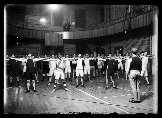 Gym class at first gym (now Barker Hall), instructor, all standing with arms outstretched