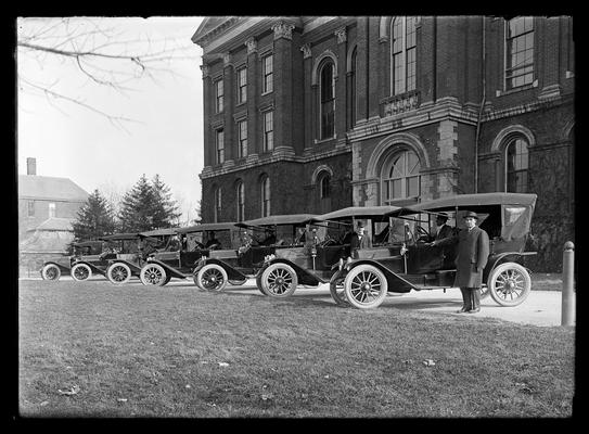 Six cars, each with driver, backed up to front of Administration Building (Main Building), one man to right of cars