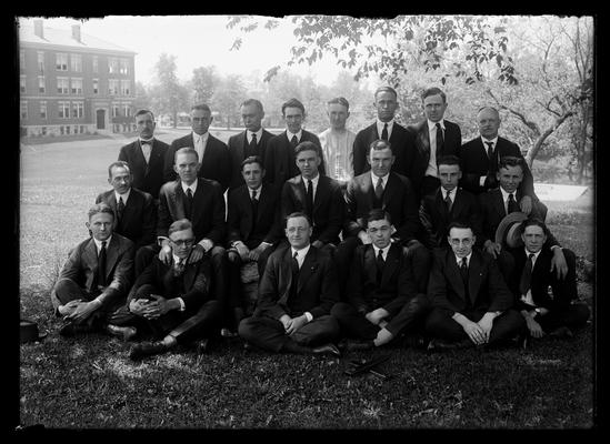 Twenty one men with Agriculture Building in background to left, one row seated, one row on knees, one row standing, left rear row is Nollau and right is Professor Anderson, two slide rules in front of group