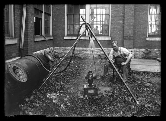 One of the Dickers outside of a building seated, in front is a lantern on a box, above is a nozzle of hose held by three crossed sticks cmoing from inside the building, Mr. Dicker is holding end of hose