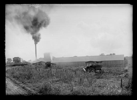 Long low building with smoke coming from smoke stack, buggy and automobile