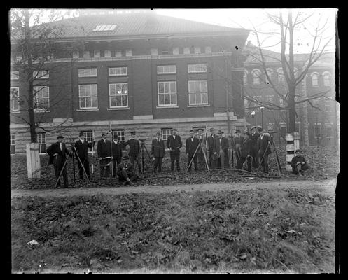 Group of men in front of building with surveying equipment, two sitting and writing