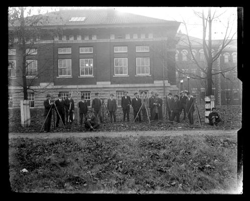 Group of men in front of building, two sitting and writing, all with surveying equipment and wearing hats