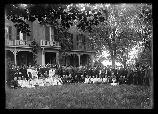 Large group of people, one row seated, all in front of brick home of director with three upstairs balconies, President Barker seated on ground near center, second from left of the men, at Scovell residence on farm