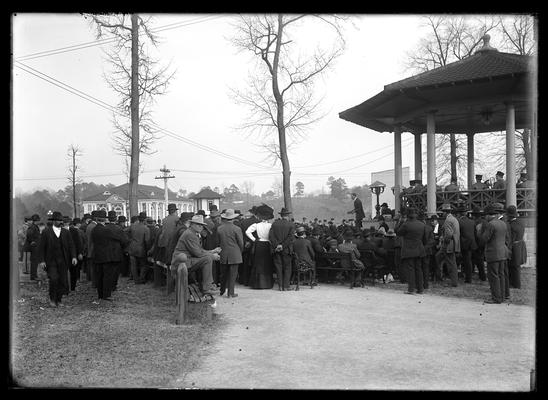 Agents meeting at Meridian, lecturing to farmers, open air pavilion to right, band, crowd seated and standing, one woman near center of picture, small boy turning to look at photographer