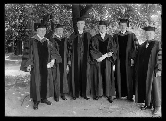 Six men in robes between Administration Building (Main Building), health building in background, F. Paul Anderson to right