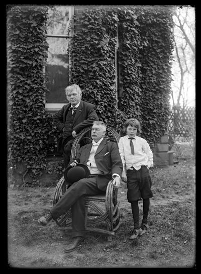 Two men and young boy, one man with mustache and derby in hand seated in chair, ivy covered building