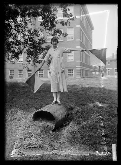 Young woman standing on barrel on side, semaphore flags, Civil Engineering and Physics Building (Pence Hall) building in background