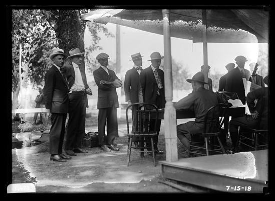 New arrivals, men with bags, others at table in front of building