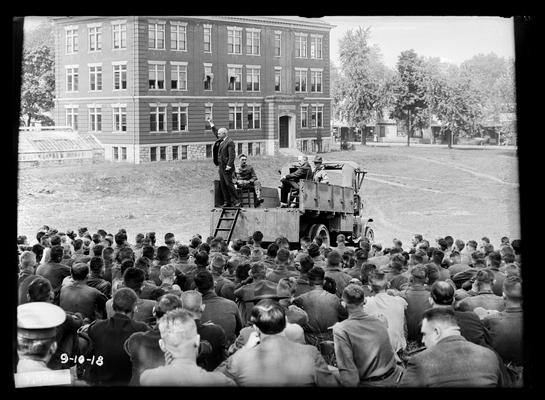 Man standing in back of truck, speaking to men seated looking toward Agriculutral building, possibly last day of camp