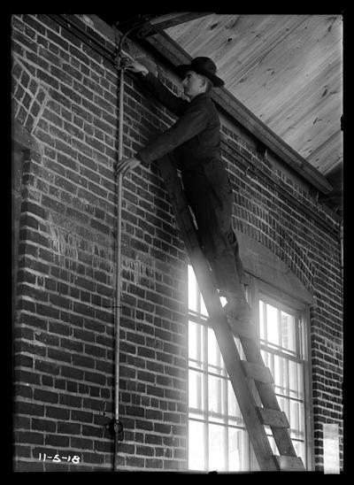 Man with hat, on ladder inside brick building, wiring conduit