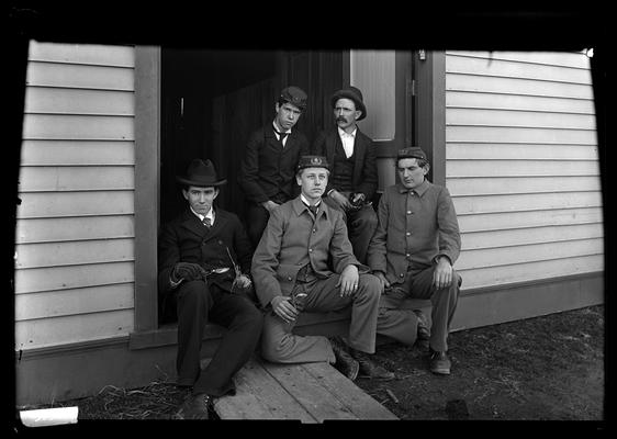 Group of five men in doorway of frame building, four have snips or tools for cutting