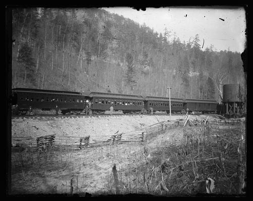 Train stopped on tracks, people standing on other set of tracks, old wooden fence