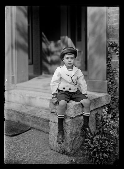 Boy in Marine suit and hat seated on stone by doorway