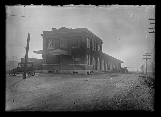 Railroad station, freight care to left, auto, bicycle, men in wagon