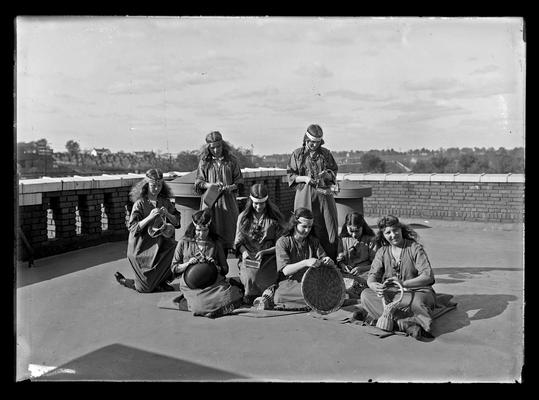 Eight girls in Indian costumes weaving baskets, on roof of Lincoln School