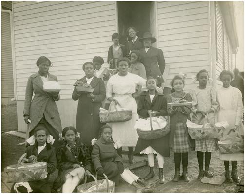 Girls holding food baskets in front of school with their teacher, Mary Graves. Handwritten on verso of 7a Brecktown Mrs. Mary Graves. (Two copies)