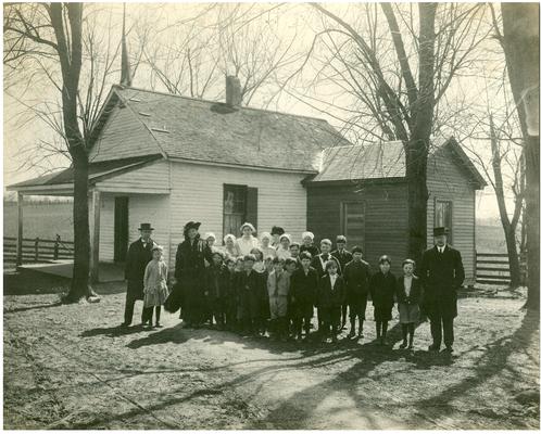 Group portrait of Locust Grove students standing in front of the school house with State School Supervisor F.C. Button, Nannie Faulconer (1865-1940), teacher Grace Yochum, and driver Mr. Davenport. Handwriting on verso (Five copies)