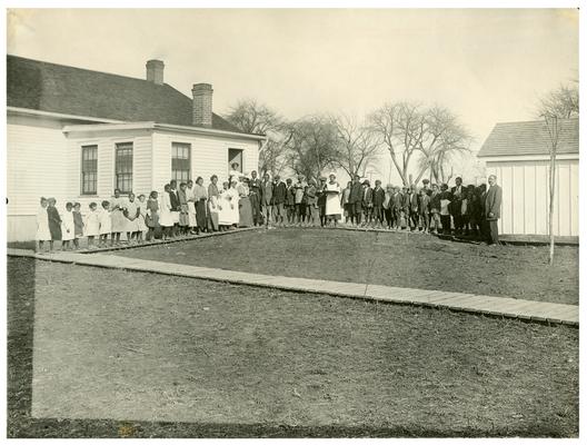 Group portrait of Maddoxtown students and Mrs. Nannie Faulconer (1865?-1940) standing in the lunch line outside of the school house. Handwritten on front, Maddoxtown-Hot lunch. Mrs. Nannie Faulconer Supt. (Four copies)